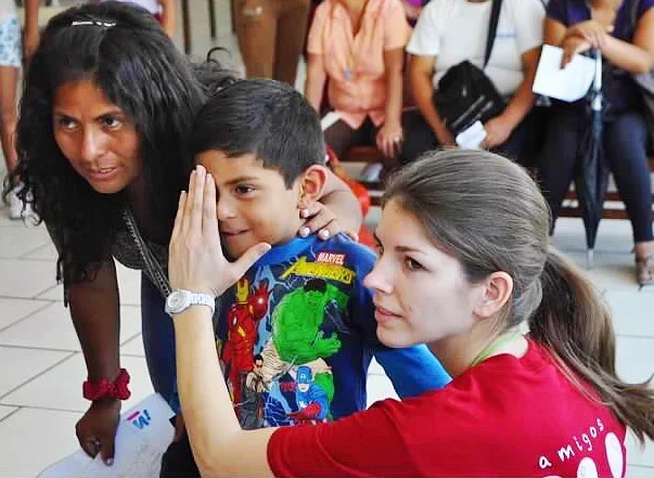 A woman with brown hair in a ponytail holds her hand to a young boy's face in a gesture of blessing or greeting, while a girl leans in closely. Several other people are in the background, some sitting and some standing, in an indoor setting.