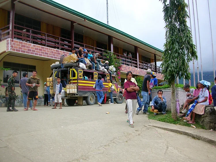 People gathered around a colorful truck with luggage on top, in front of a two-story building with a balcony and large windows. Some people are sitting on the truck, while others are standing or sitting on rocks and benches nearby. There is a tree on the right side and mountains in the background.