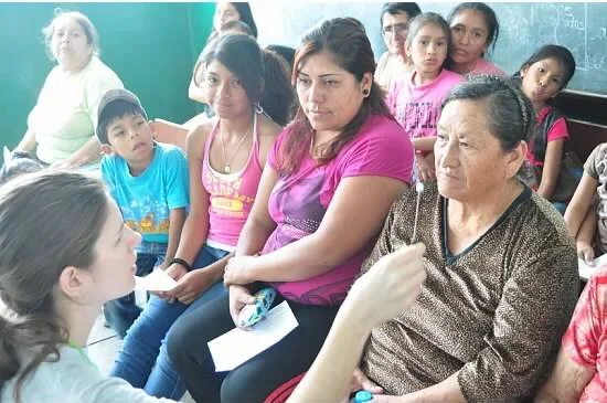 A group of people, including children and women, sitting and listening to a woman in a classroom.