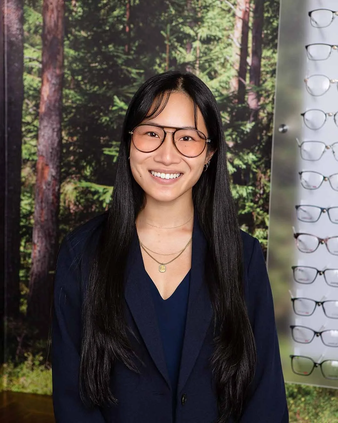 A woman with long black hair, wearing glasses, smiling, dressed in dark blue blazer and top, standing in front of a forest-themed backdrop with sunglasses display on the side.