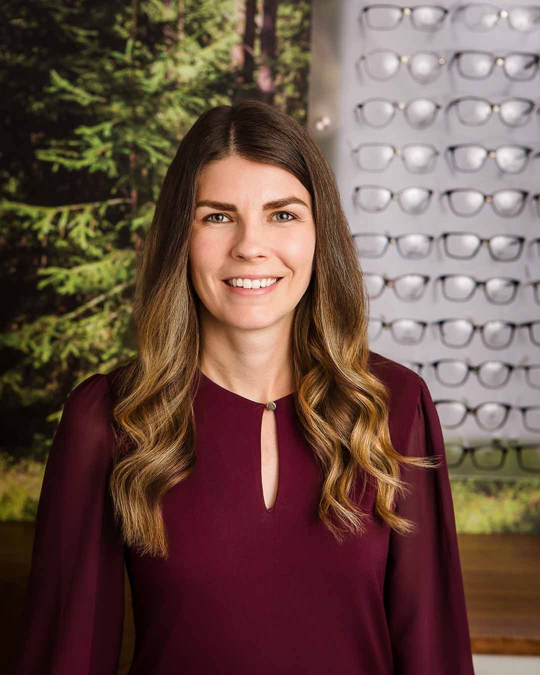 A woman with long wavy brown hair smiling in an eye care store, with glasses displayed on a wall behind her.
