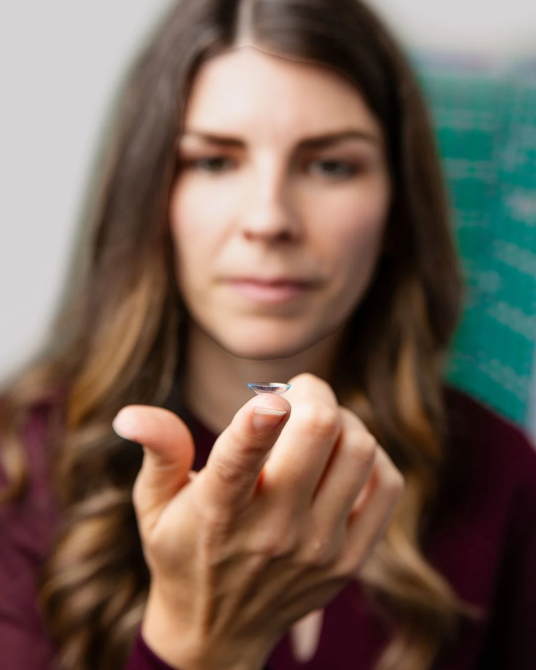 A woman with brown hair holding a contact lens on her finger.