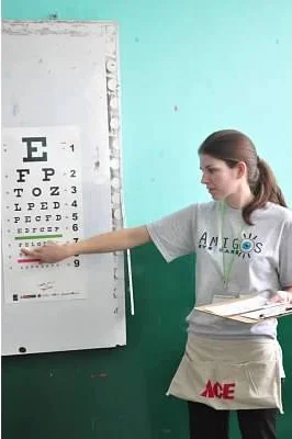 A young woman standing in front of an eye chart on a whiteboard, pointing at the chart, and holding a notebook, inside a classroom.