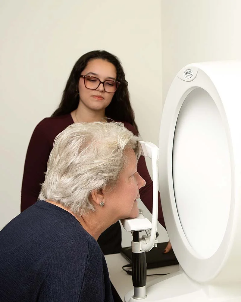 A woman undergoing a medical imaging procedure with her head positioned in a machine as a technician observes.