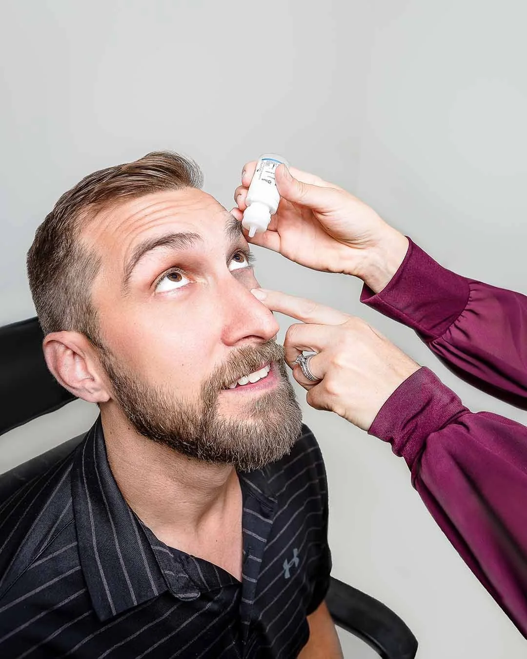 A man with a beard and mustache sitting in a chair, looking upward as a woman applies eye drops to his left eye with a small dropper bottle.