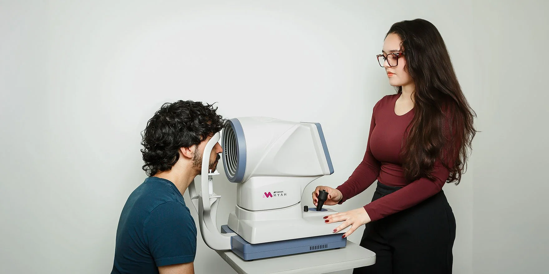 A woman is performing an eye test with an ophthalmic device at the eye clinic, while an optometrist or technician observes and operates the equipment.