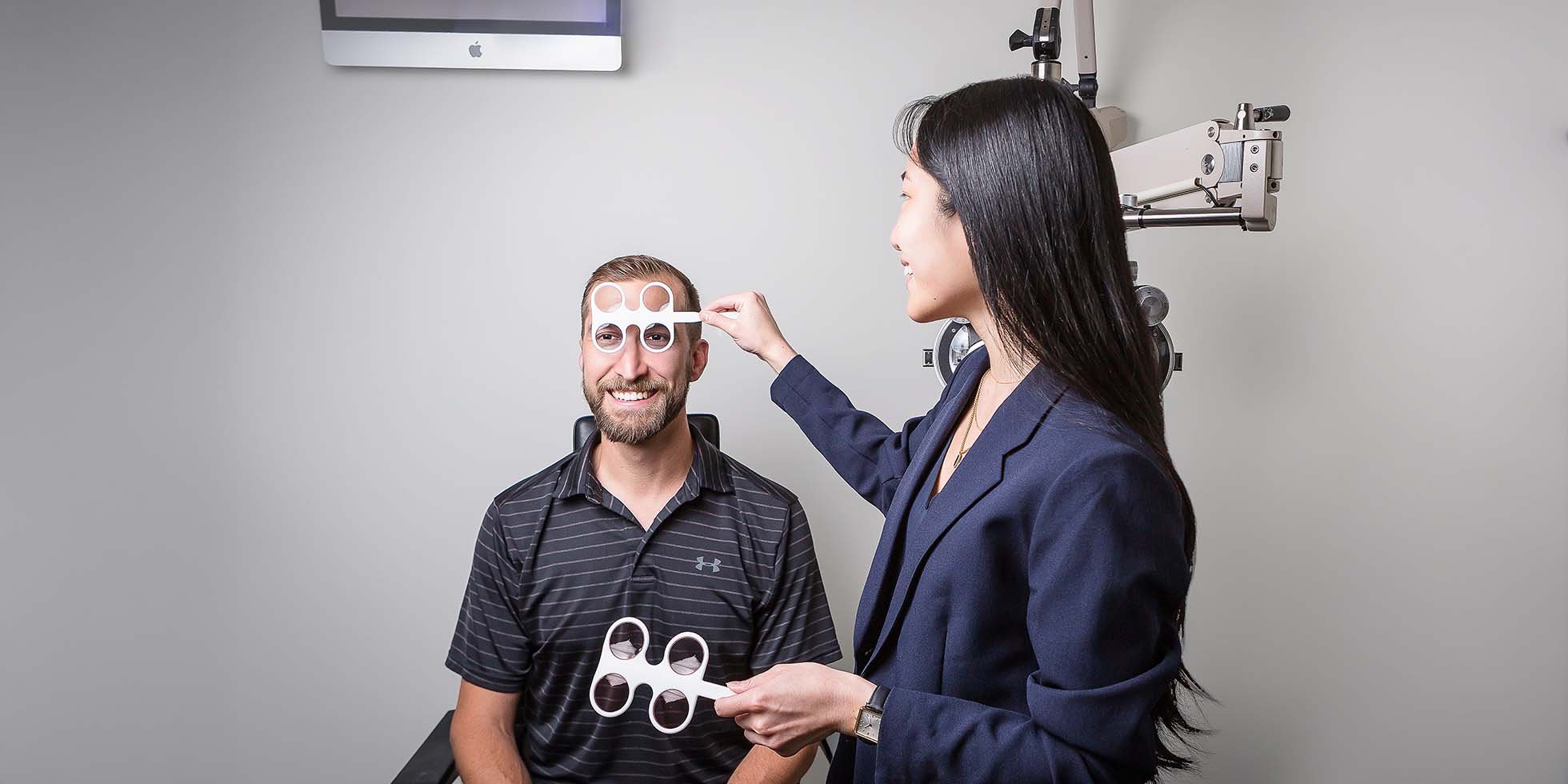 A woman conducting an eye exam on a man using a phoropter with trial frames nearby in an ophthalmologist's office.