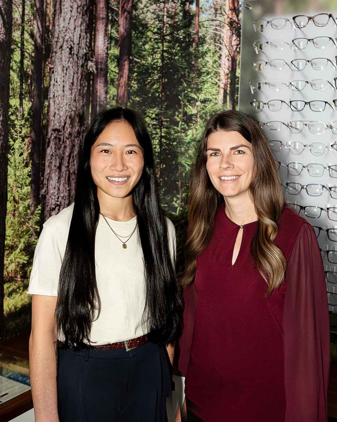 Two women standing indoors, one wearing a white shirt and black pants, the other wearing a burgundy dress, with a trees and forest background and display of eyeglasses behind them.