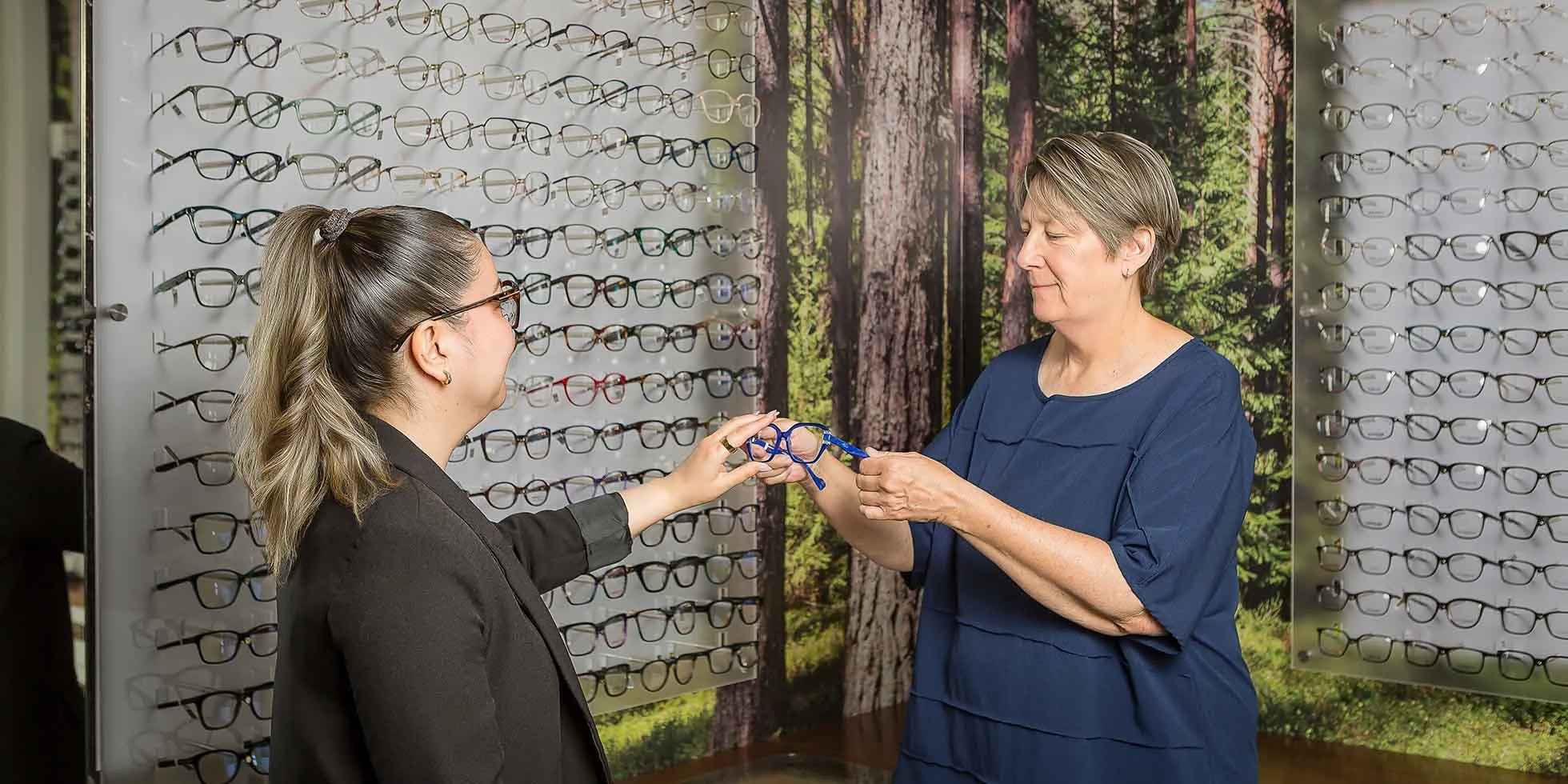 A customer is trying on glasses with the assistance of an optician or store employee at an eyewear store or optical shop, with shelves of eyeglasses displayed on the wall behind them.