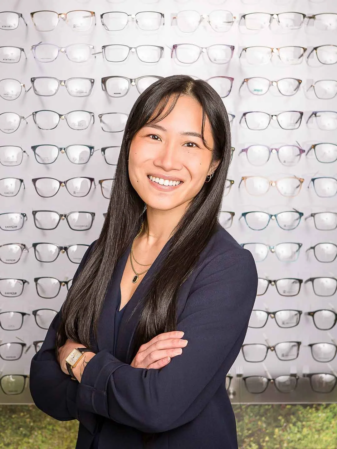 A young woman with long black hair and a bright smile standing with her arms crossed in front of a display wall filled with various eyeglasses.