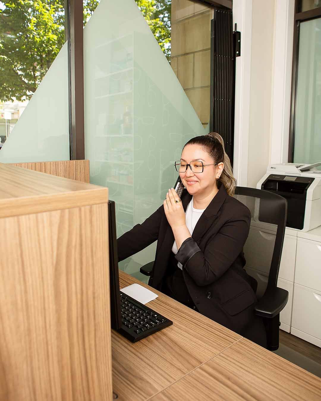 A woman in a business suit talking on a landline phone at her desk in an office with large windows and green trees outside.
