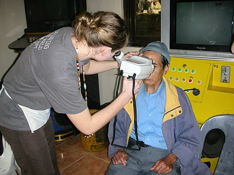 A healthcare worker or caregiver uses a diagnostic device to examine an elderly man sitting in a room with medical equipment.