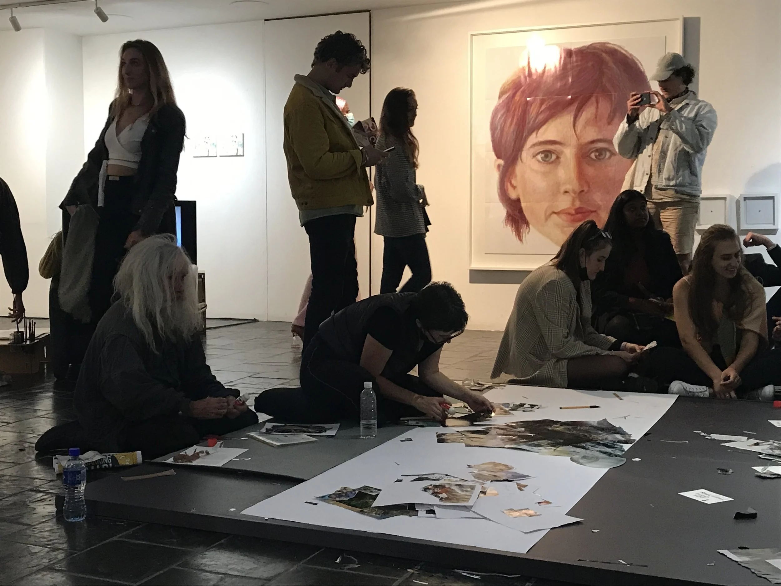Mensen in een galerie kijken en maken kunst, met grote portretkunstwerk op muur, tafels met kunstmaterialen en mensen die werken en fotograferen.