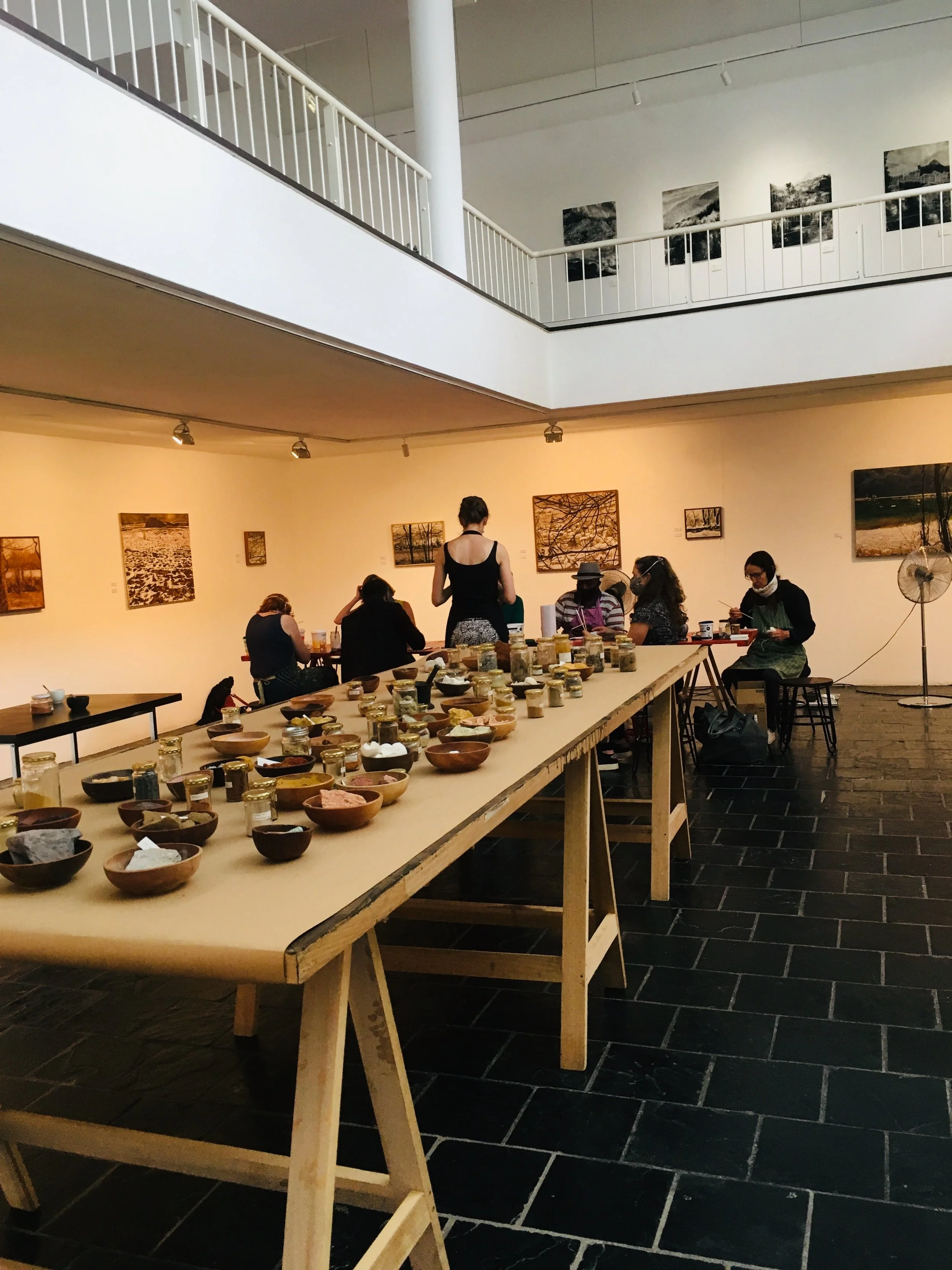 Binnen in een kunstgalerij of studio met mensen die aan een tafel werken of voor zich uit kijken, en veel aardewerken kommen en potten op een grote tafel.