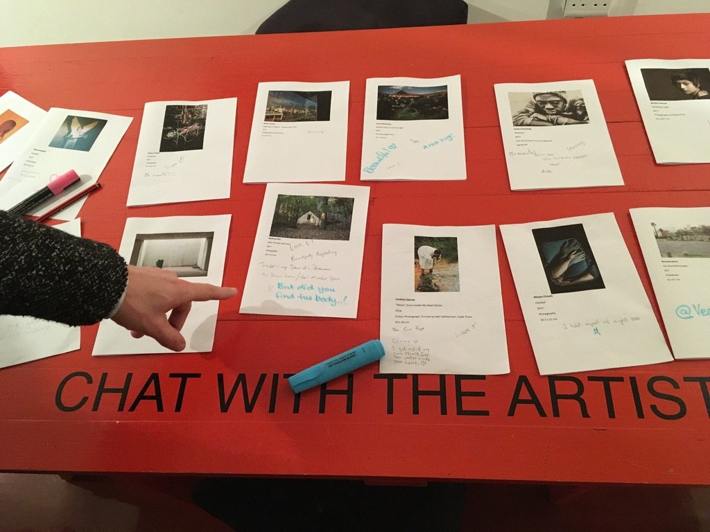 Fotolijsten met foto's en notities op een rode tafel met de tekst 'CHAT WITH THE ARTIST'.