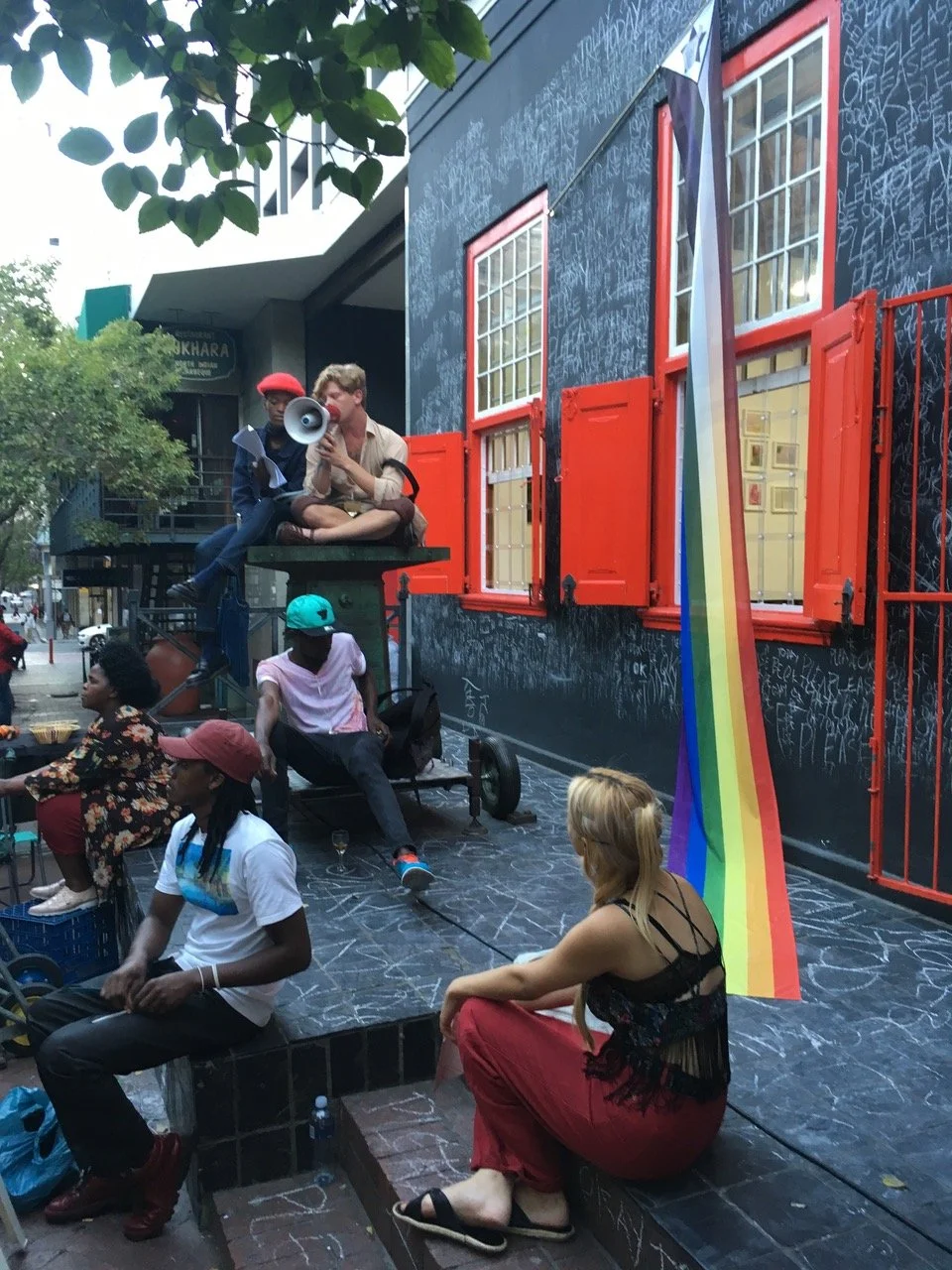 Diversen mensen zitten en staan buiten voor een muur met graffiti en een regenboogvlag. Een man met een megafoon en een vrouw zitten boven op een platform, terwijl anderen op de grond zitten of staan.