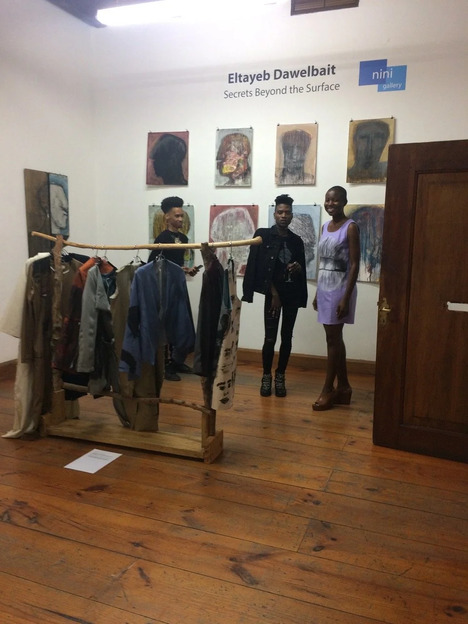 Tentoonstelling met kunstwerken van Eltayeb Dawelbait, getiteld 'Secrets Beyond the Surface', met drie mensen die in de galerie staan, en kledingstukken op een houten rek.