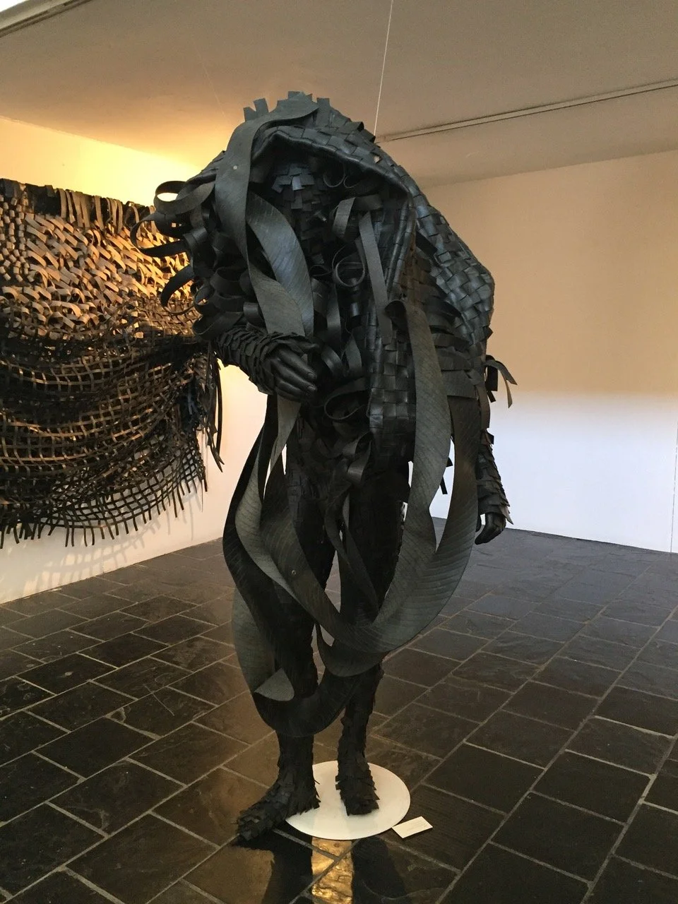 Een zwart sculptuur van een menselijk lichaam gemaakt van bandjes en stroken van materiaal, getoond in een kunstgalerie.