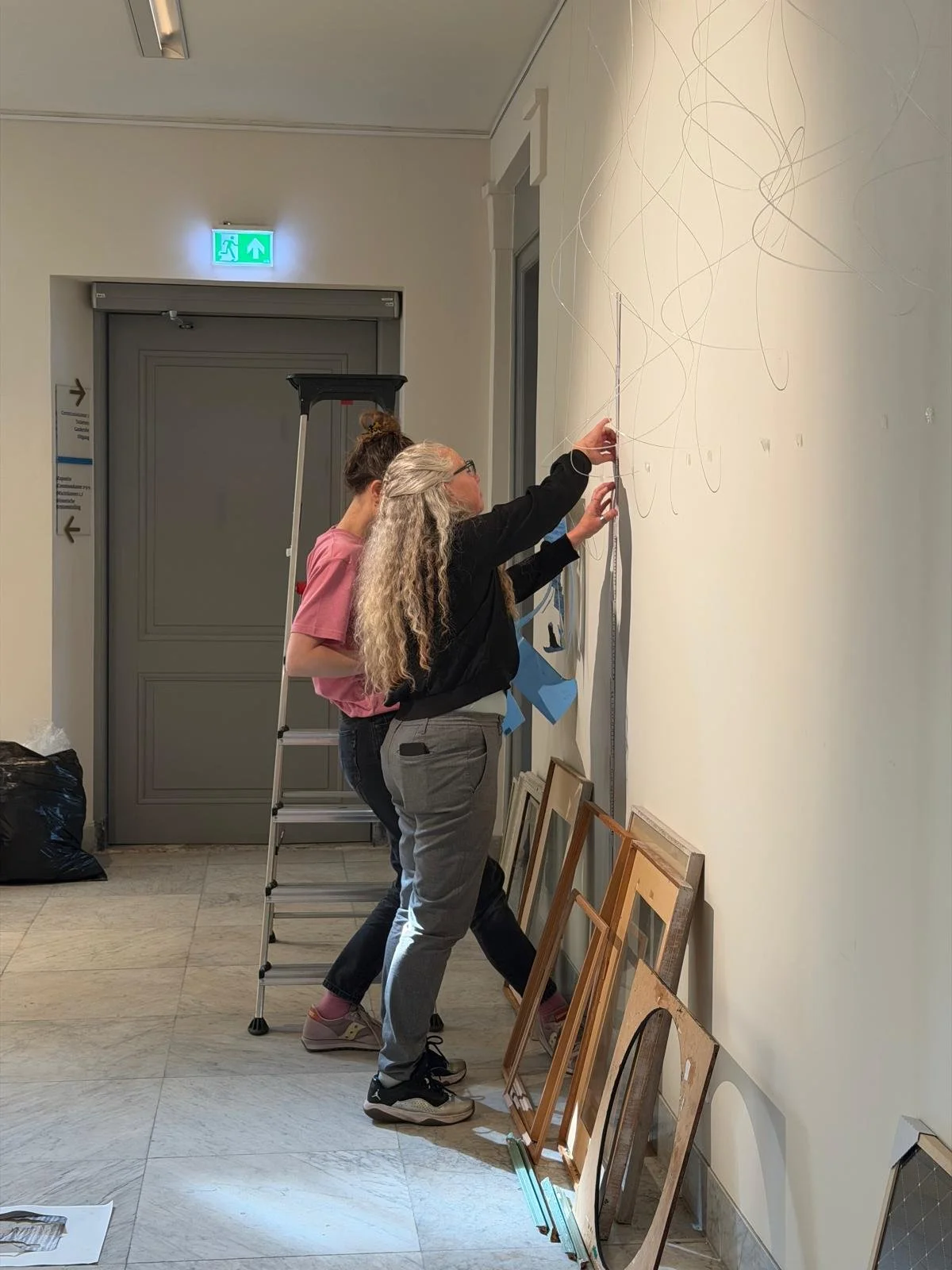 Twee vrouwen hangen schilderijen op aan een muur in een galerij of interieurruimte.