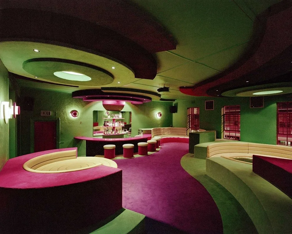 Leeg interieur van een bar met groene en roze kleuren, ronde zitplaatsen en een bar met drankjes.