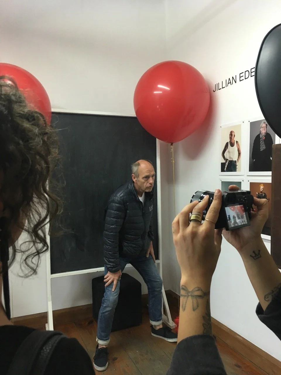 Man poseert in een gallery met foto's aan de muur en rode ballonnen, terwijl iemand foto's van hem maakt.