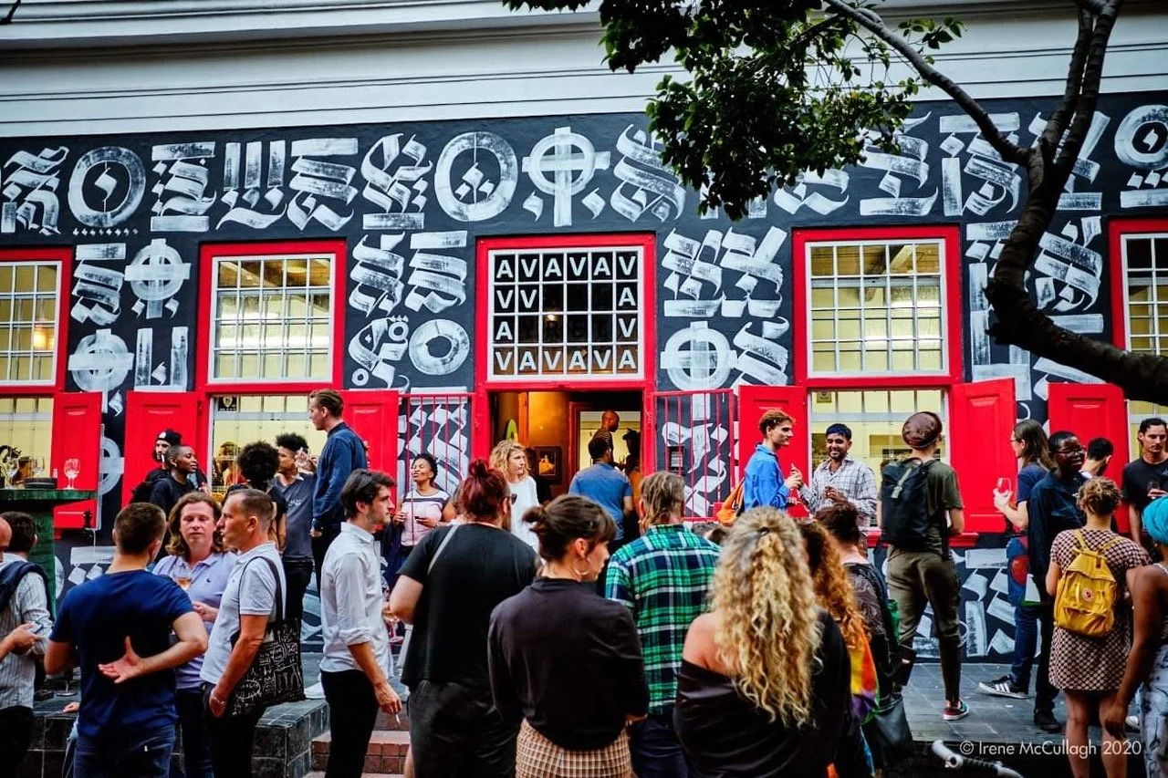 Een drukke buitenbar met mensen die praten en drinken, voor een restaurant of café met een graffiti-achtige muurschildering en rode verkiezingramen.
