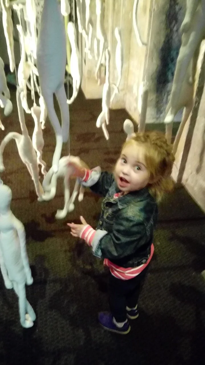 Een jonge kind met een denim jas en een roze en witte gestreepte trui, die naar witte flamingo-beeldjes op een tentoonstelling of in een museum kijkt en ze aanraakt.
