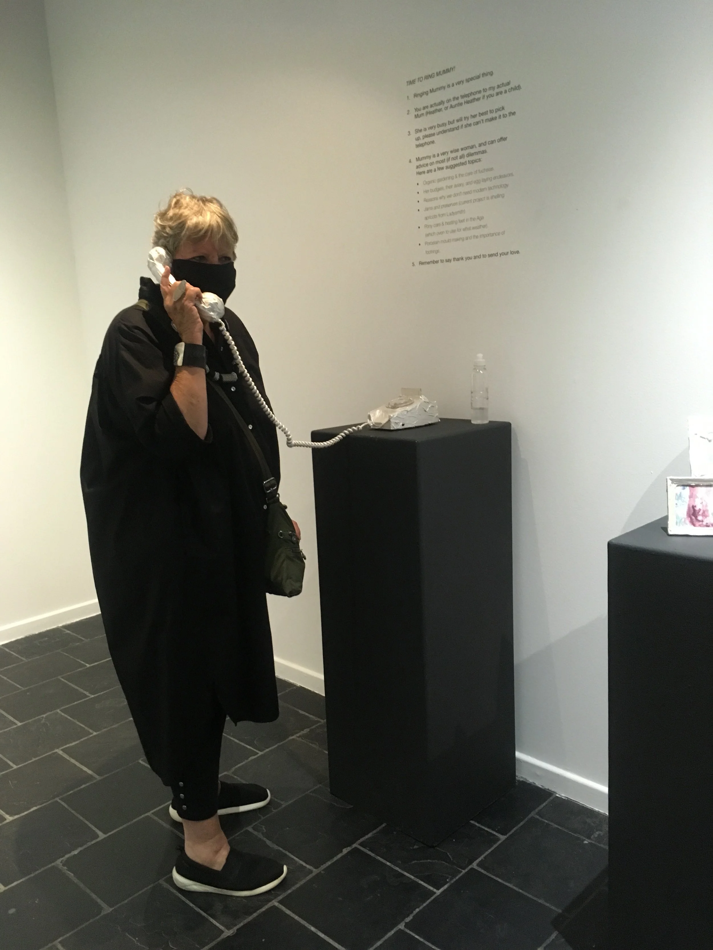 Een vrouw met een zwart mondkapje en zwarte kleding die een oud telefoontoestel vasthoudt in een kunsttentoonstelling. Ze staat op een zwarte tegelvloer naast een zwarte sokkel met water en andere objecten erop.