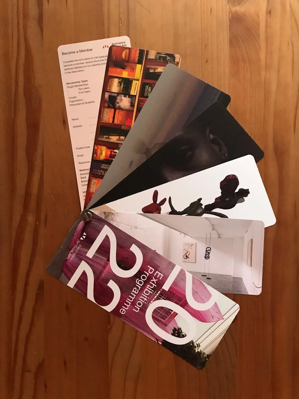 Verschillende brochures en catalogi liggen uitgevouwen op een houten tafel, inclusief een brochure over kunstuitingen en een brochure van 125 jaar, naast een paar kaarten met afbeeldingen van een kunstwerk en een gebouw.