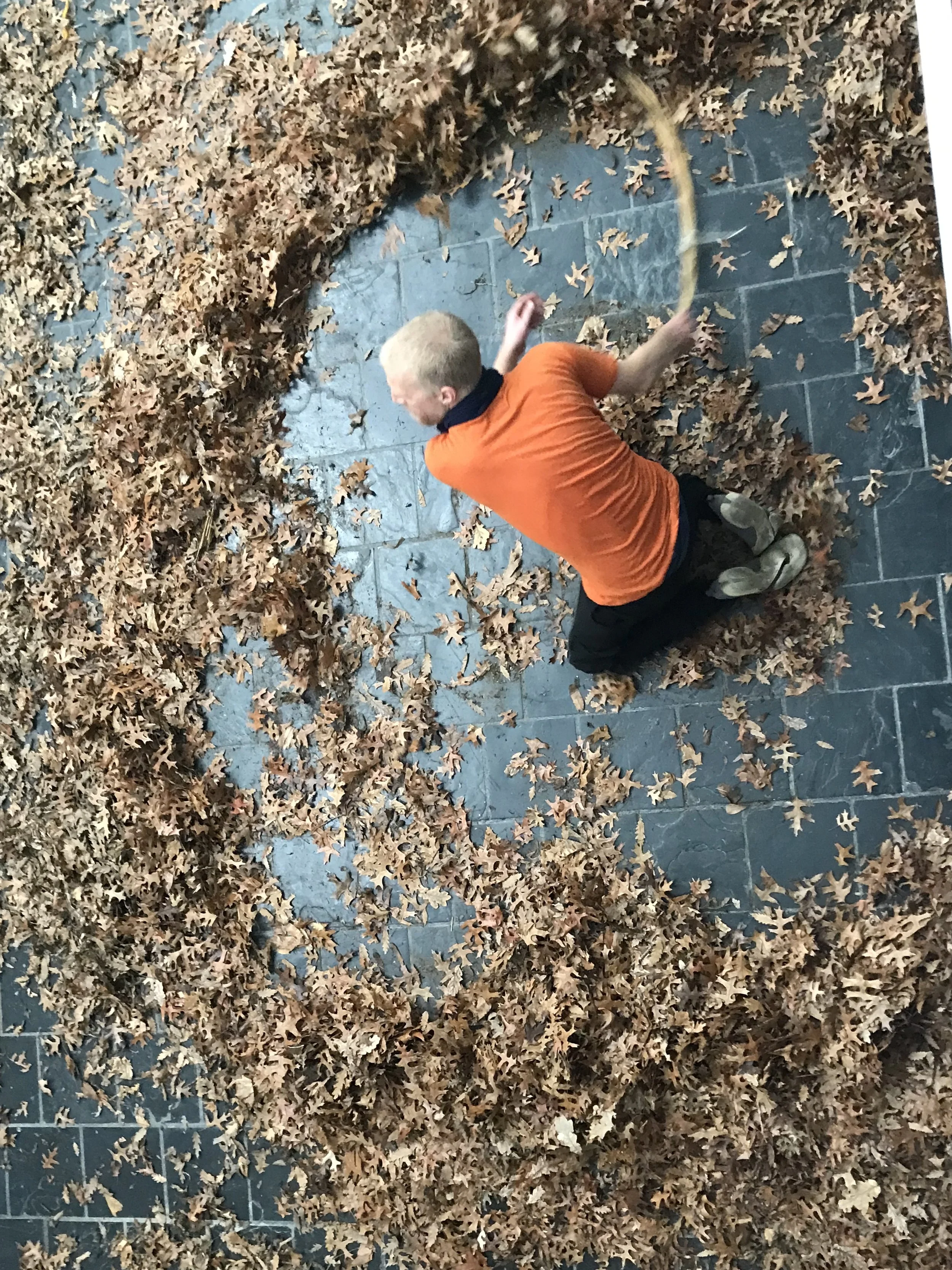 Een jongen ligt op de grond omgeven door een grote hoeveelheid uitgewaaide herfstbladeren buiten op een tegelvloer.
