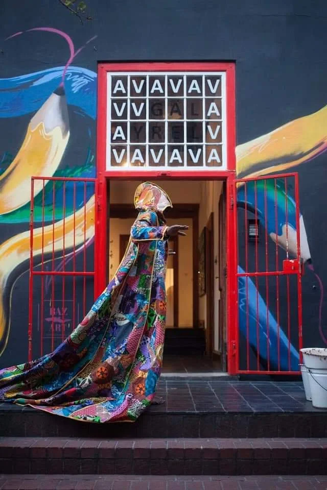 Een kleurrijk kunstwerk met een muze-achtig standbeeld dat een patchwork jurk draagt, staande voor een muur met kleurrijke muurschilderingen van een penseel en abstracte vormen, en een bord met gekleurde blokken met letters.
