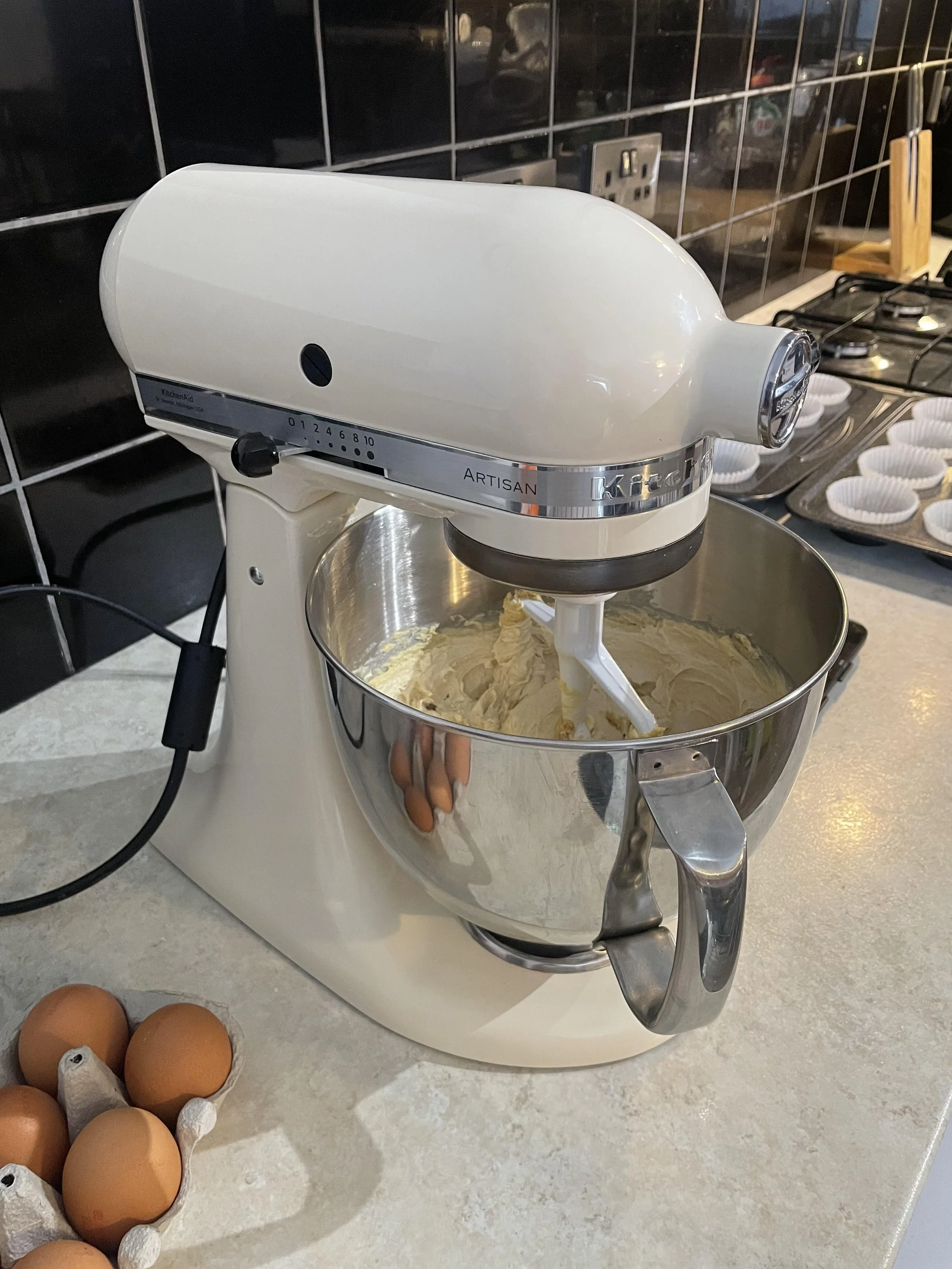 A white kitchen stand mixer with a metal mixing bowl, mixing batter or dough, on a kitchen countertop. There are eggs in a carton nearby and cupcake liners on a tray in the background.