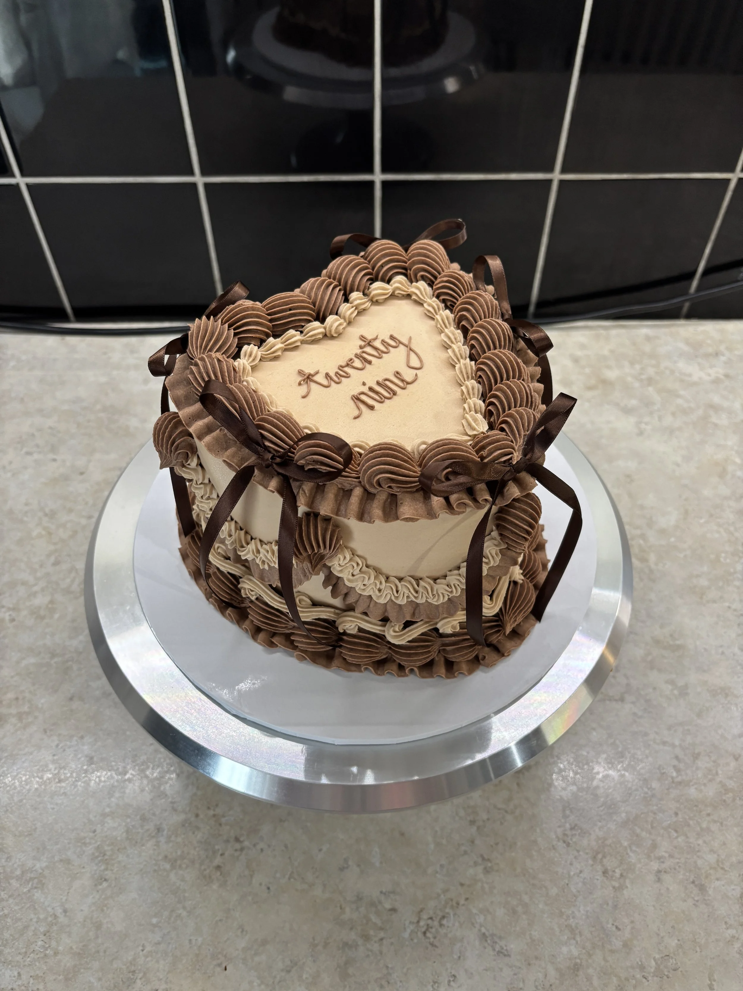 A heart-shaped birthday cake with cream and chocolate frosting, decorated with chocolate ribbons and swirls, on a silver cake stand.