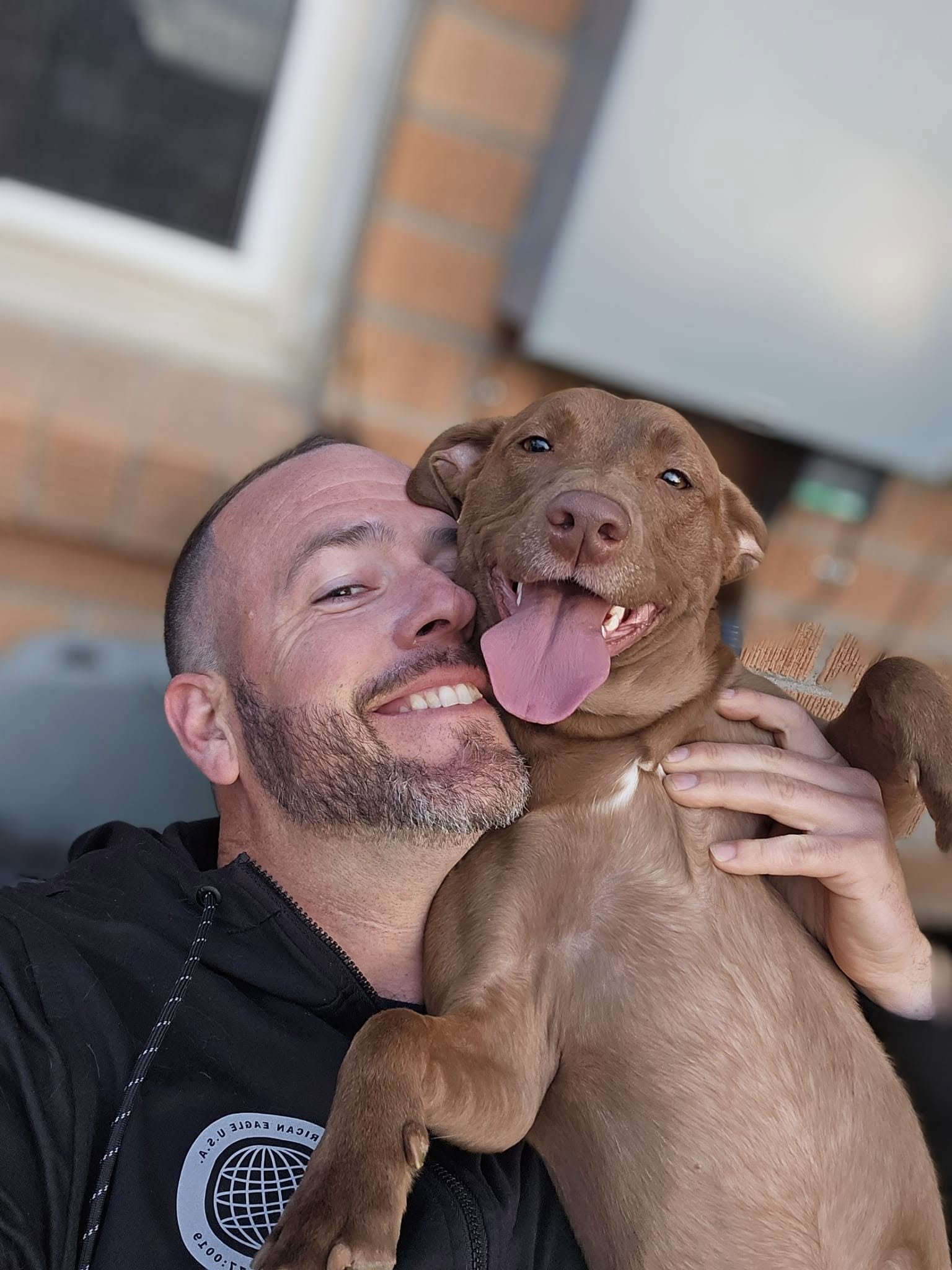A man smiling and hugging a large brown dog with its tongue out, in front of a brick wall.