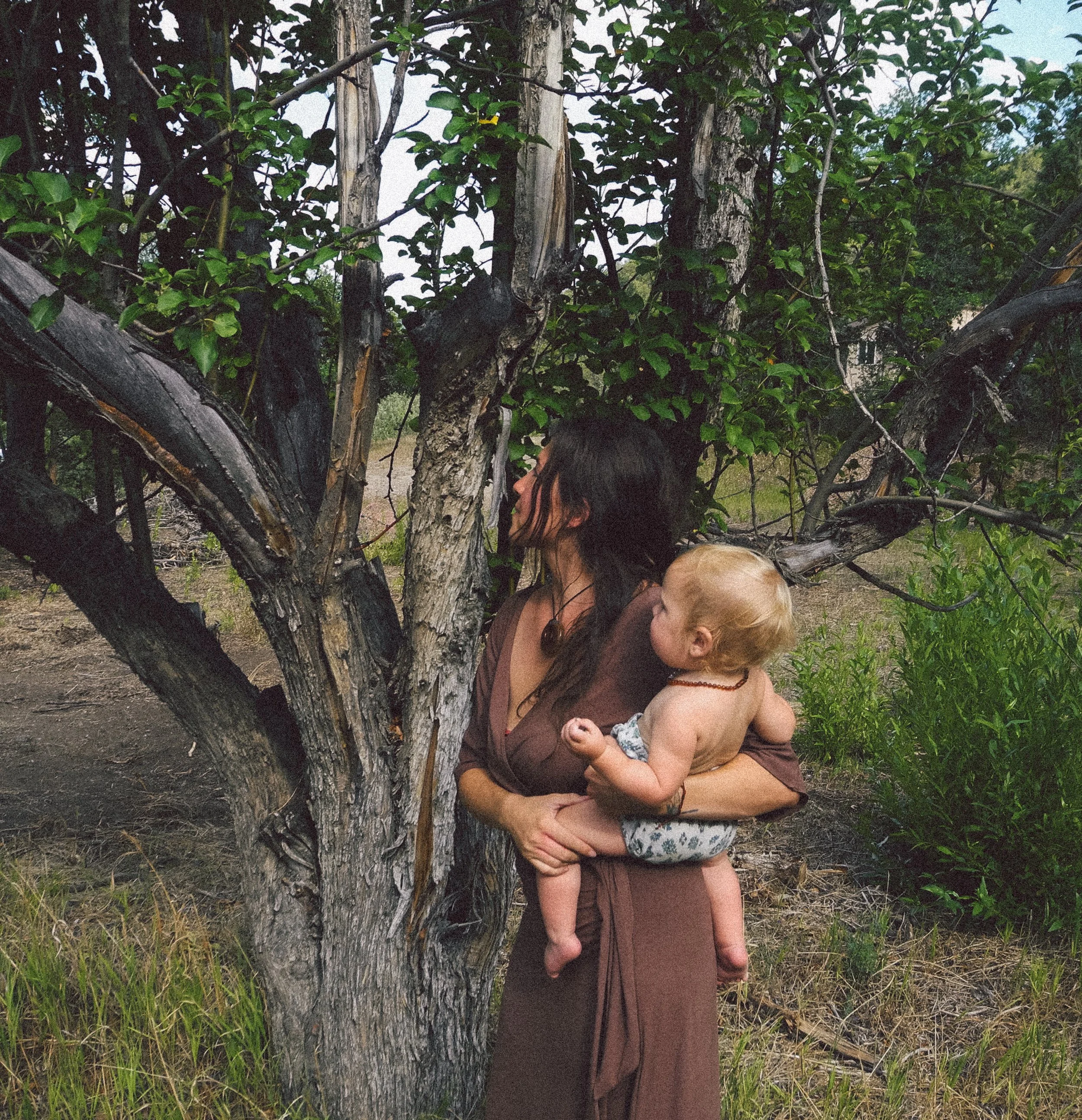 A woman holding a child stands in front of a large tree, surrounded by green grass and foliage, with both of them partially obscured by the tree's branches.