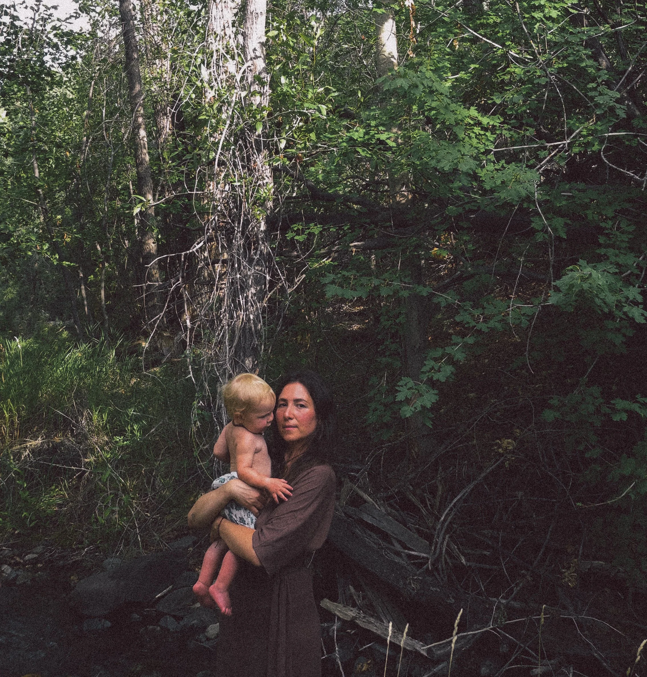 A woman holding a young child in her arms standing in a forested area near a small stream or river.