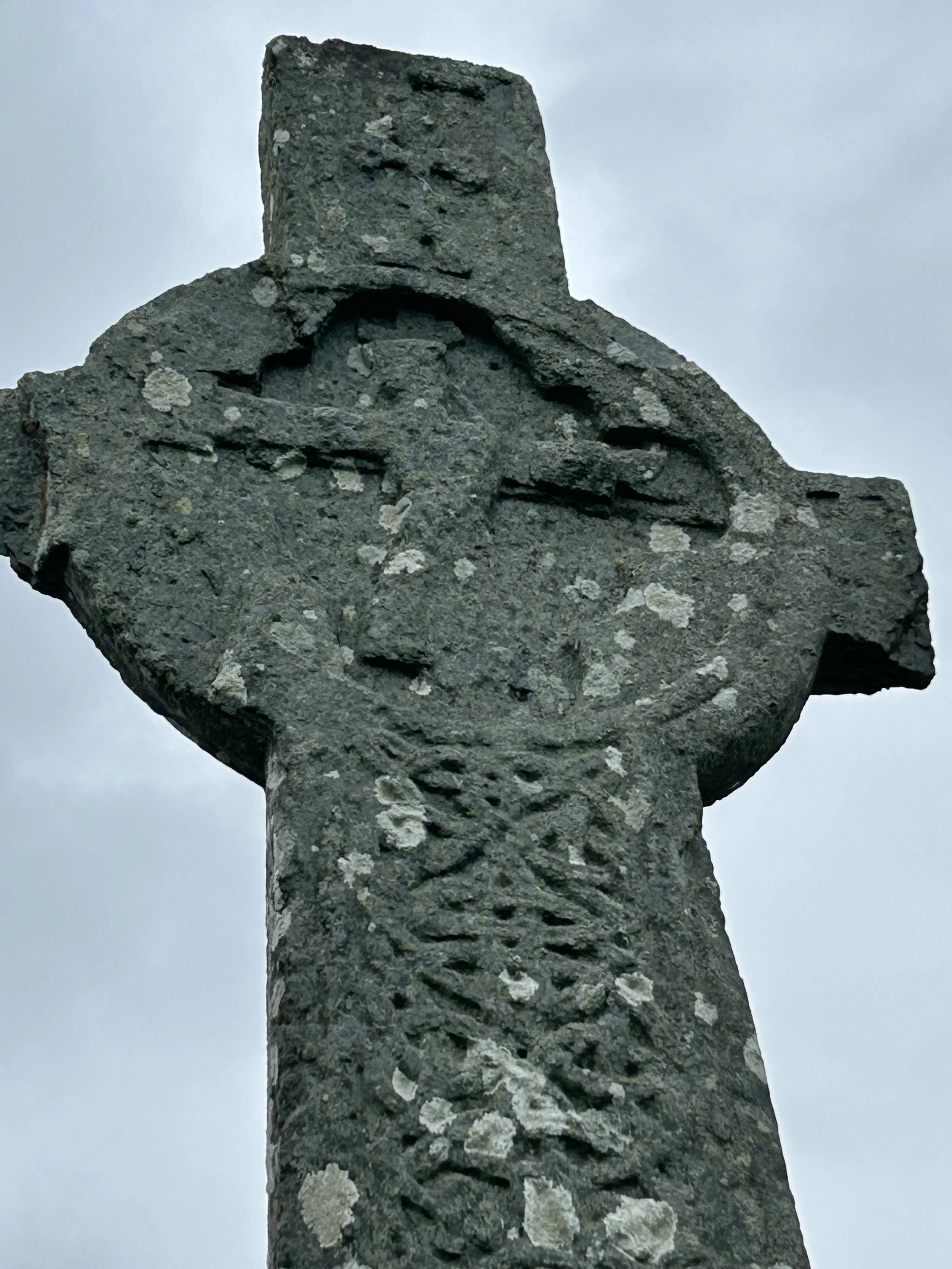 Iona, Scotland: St Martin's Cross, which has stood on its spot for over 1200 years. Close-up of a weathered, ancient stone cross with a carved Mary and Jesus in the center, against a cloudy sky background.