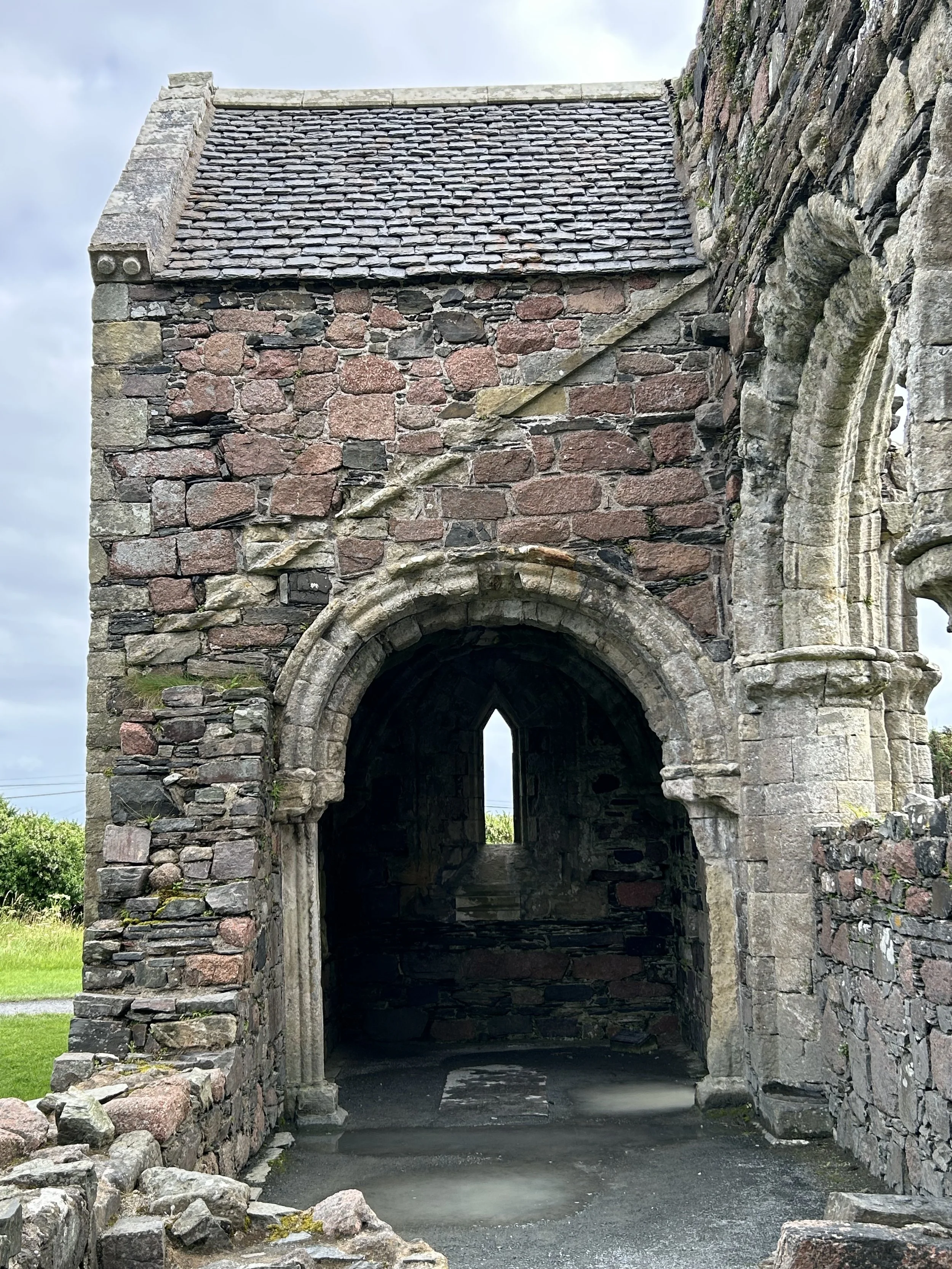 Iona, Scotland: The Nunnery featuring an ancient stone building with a pointed arch doorway and a small window of light and a vaulted ceiling, made of large rough-cut stones and bricks, with a roof covered in slate tiles.