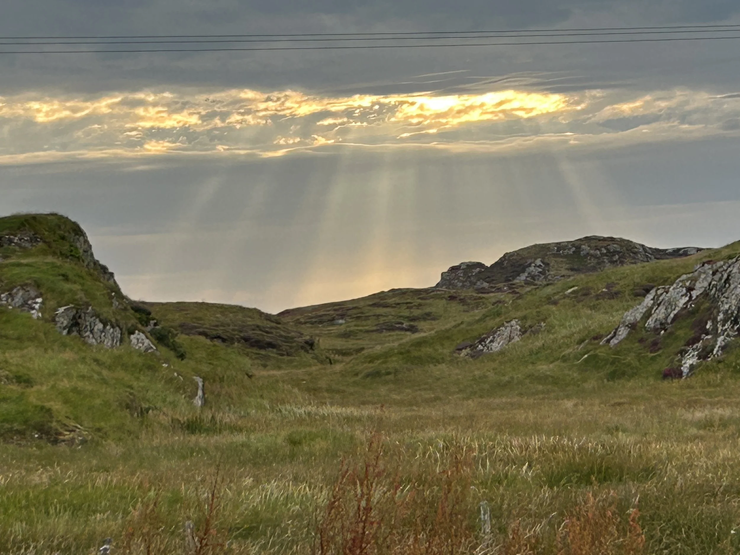 Iona, Scotland: A scenic landscape of rolling green hills with rocky outcroppings, under a sky with partly cloudy conditions and sun rays breaking through, creating a serene view.