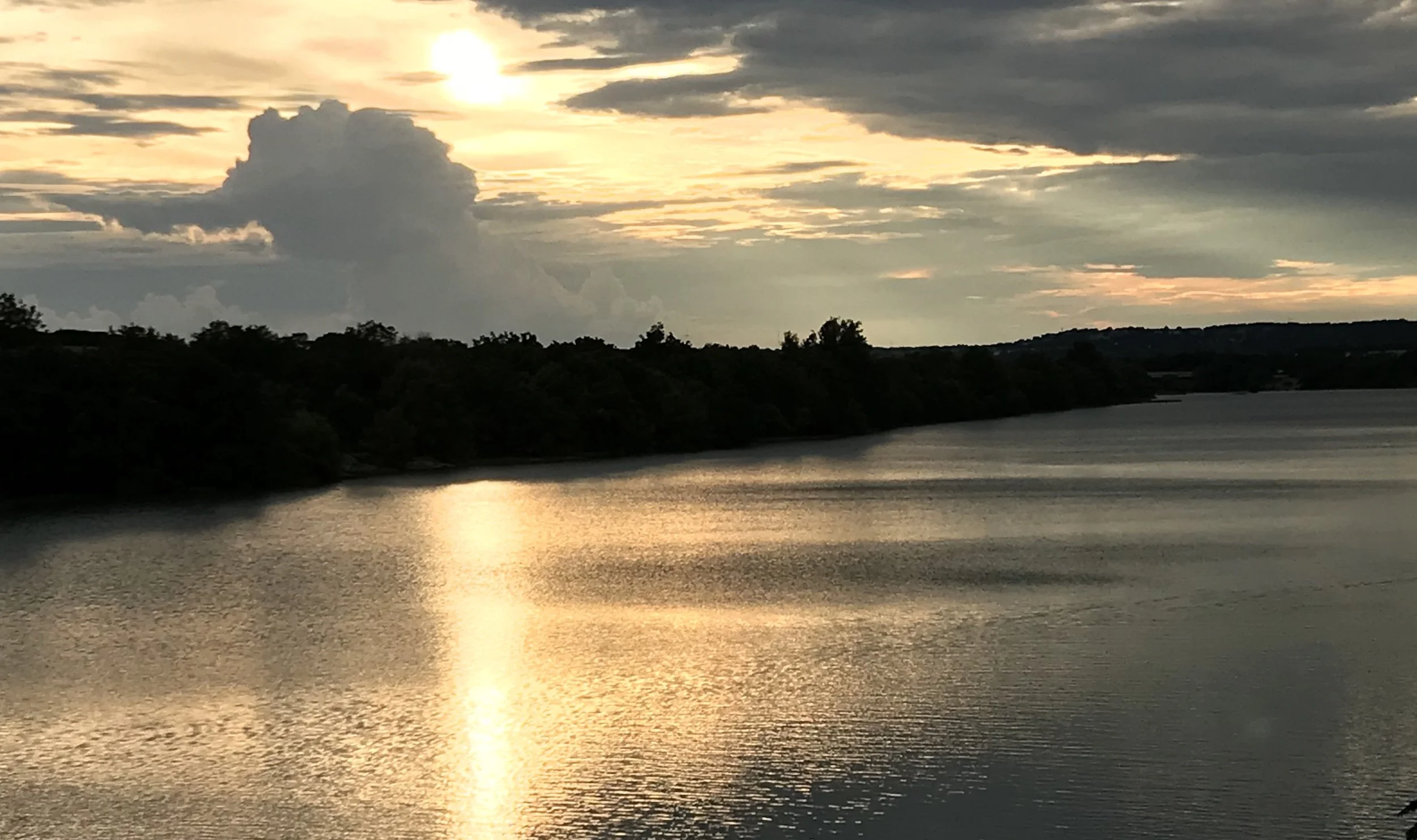 Kerrville, TX: Sunset over the Guadalupe river with the reflection of the sky and clouds on the water surface, featuring a mix of dark and illuminated clouds.