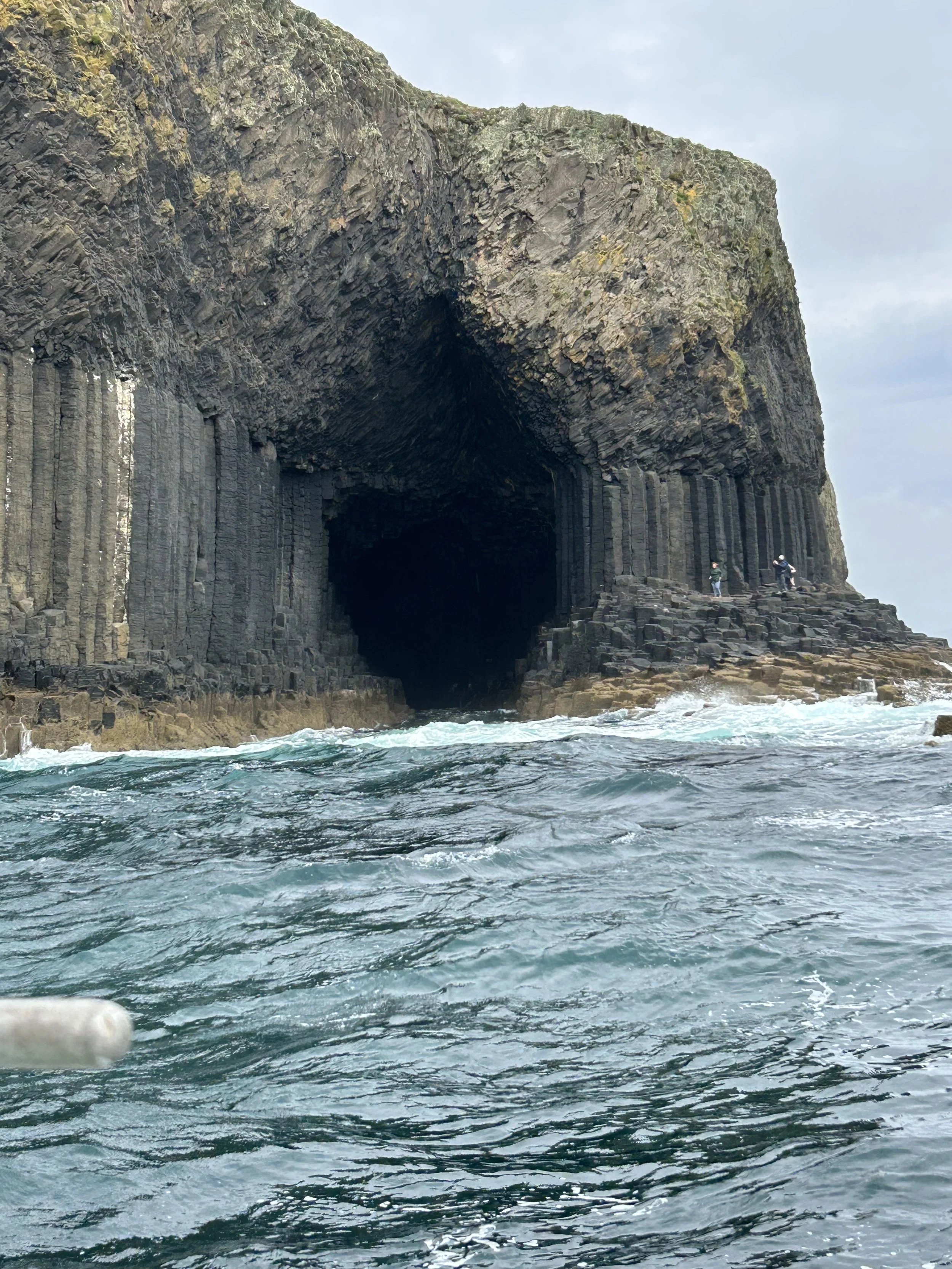 Hebrides Islands, Scotland: Cliffside of Fingal's Cave, rocky formations, and people walking on rocks near the ocean.