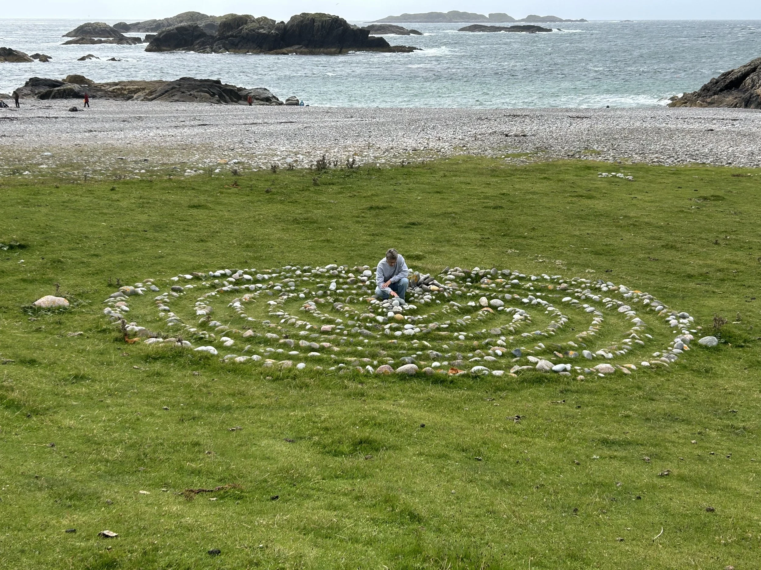 Iona, Scotland: Labyrinth at Columba's Bay - A person in the center of a pebble labyrinth near a beach with rocks and ocean in the background, cloudy sky.