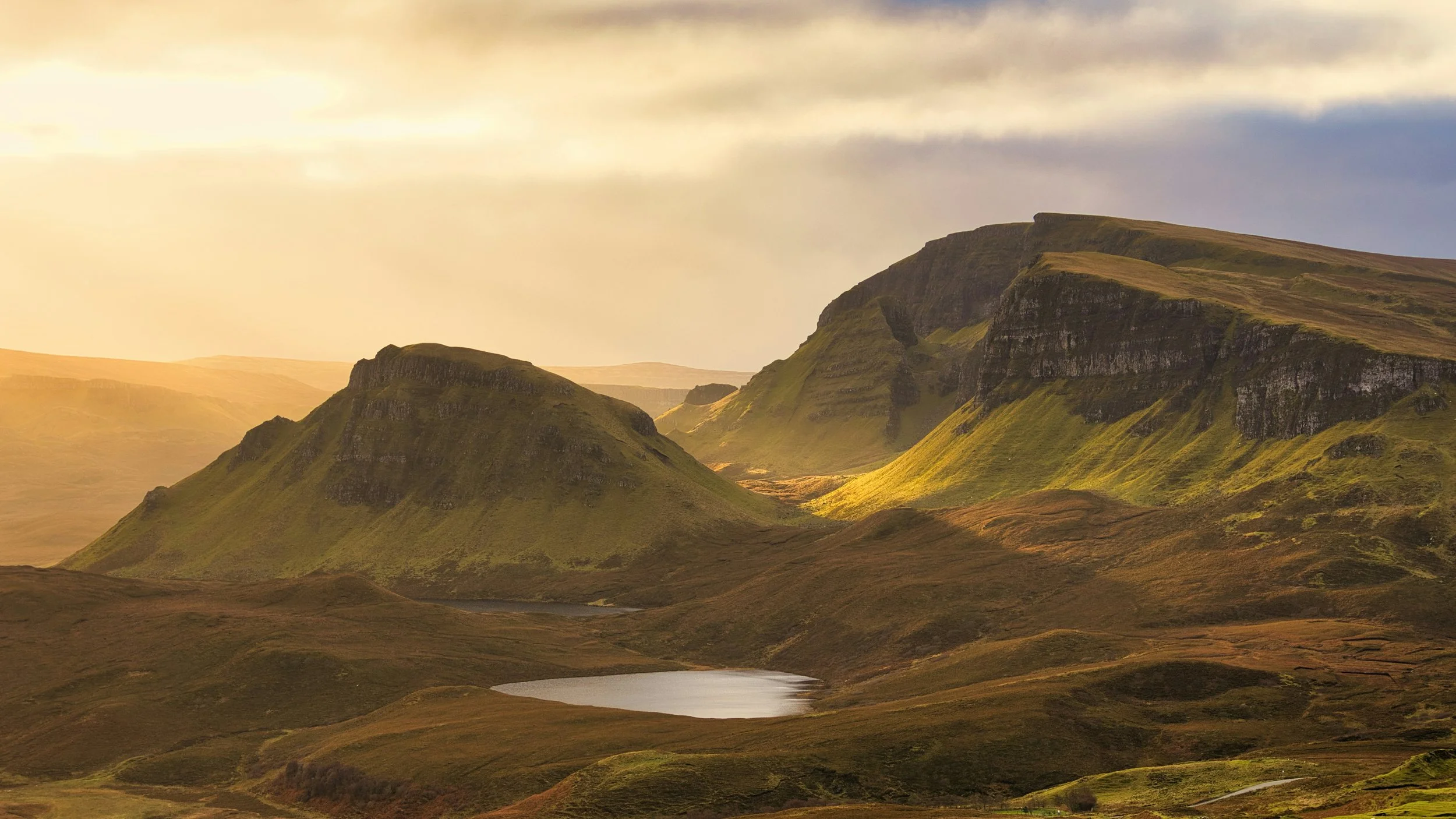 Sunlight illuminating green hills and rugged cliffs over a small lake in a mountainous landscape.