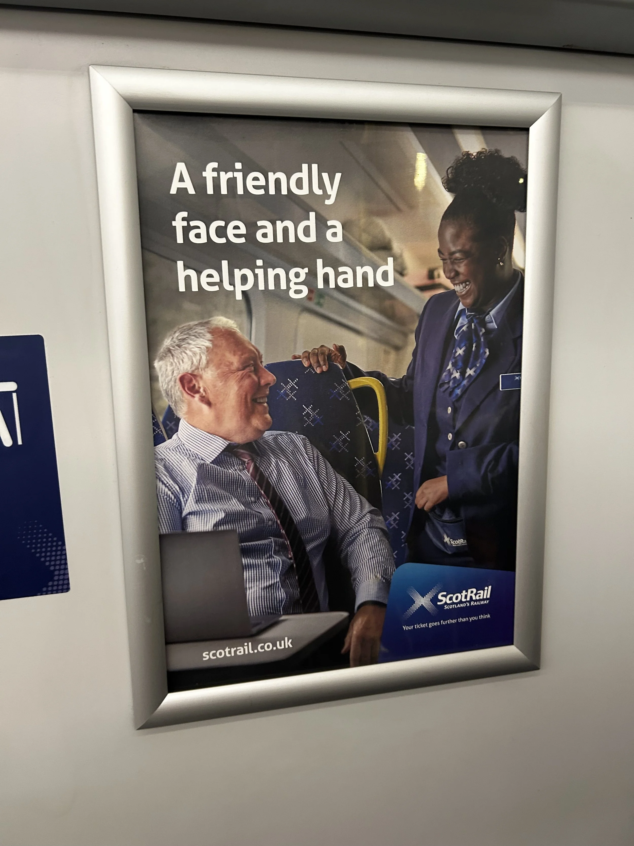 An advertisement poster on a train or bus featuring a smiling man and woman, with the text 'A friendly face and a helping hand' and the logo of ScotRail, Scotland's Railway, at the bottom.