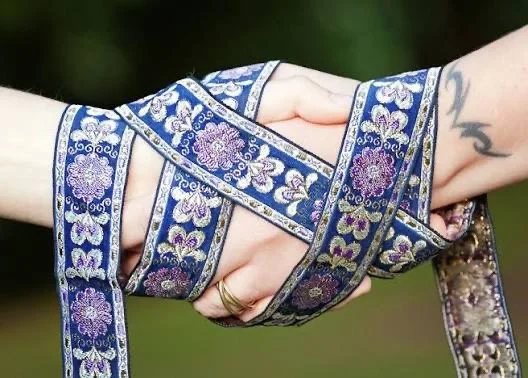 Two people holding hands, intertwined with a decorative blue and purple embroidered ribbon.
