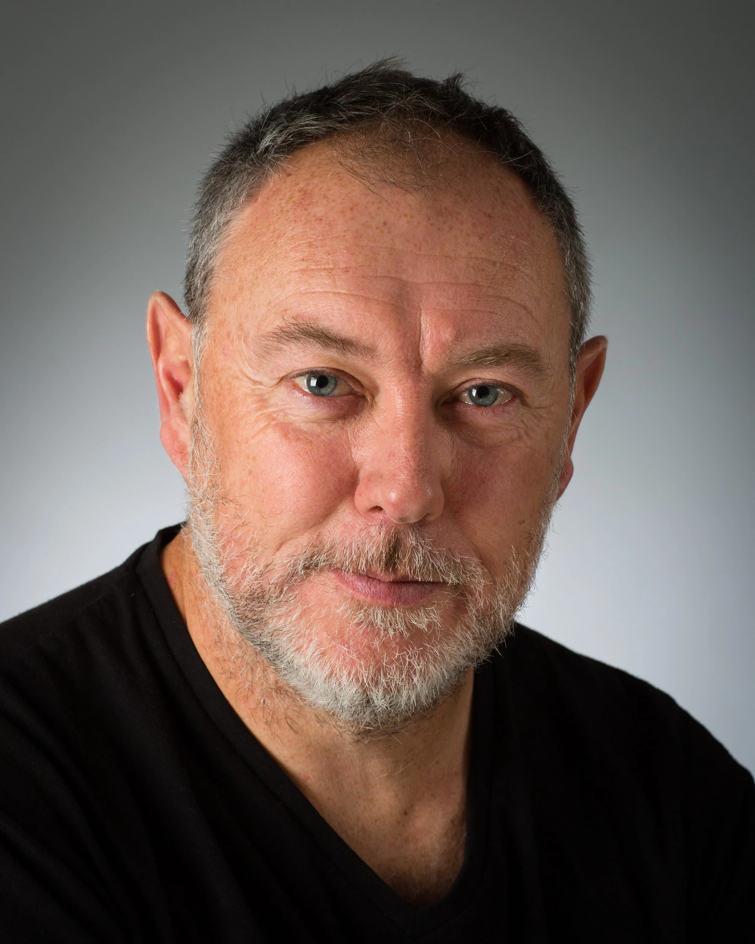 Close-up portrait of mature man with blue eyes, gray beard, and short gray hair, wearing a black shirt against a neutral background.