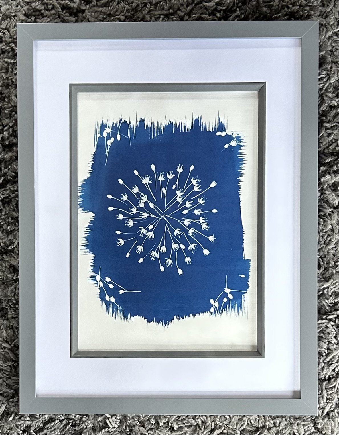 Framed abstract art with blue and white colors, resembling a floral or botanical design, placed on a gray textured surface.