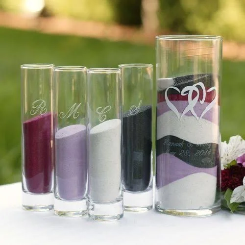 Five pink, purple, white, black, and multi-colored candles or decorative glasses with carved initials and designs, set outdoors on a white surface.
