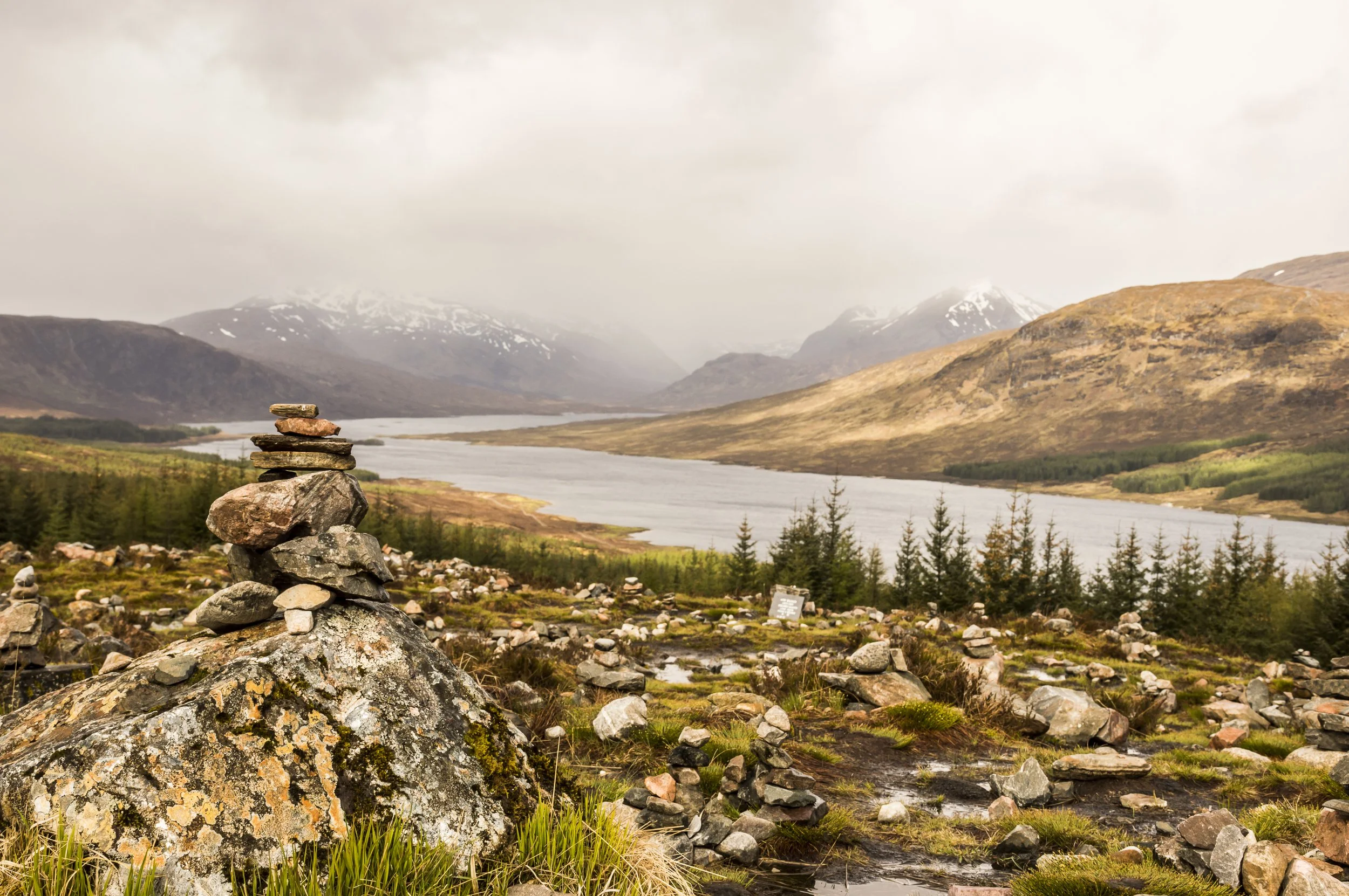 A landscape with a rocky foreground, small cairn of stacked stones, a river extending into the distance, and mountains with snow patches under cloudy skies in the background.