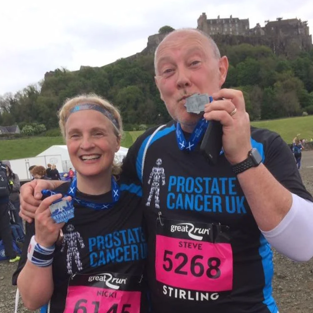 Two runners in Prostate Cancer UK running shirts celebrating after completing a race, holding medals, with a historic castle on a hill in the background.